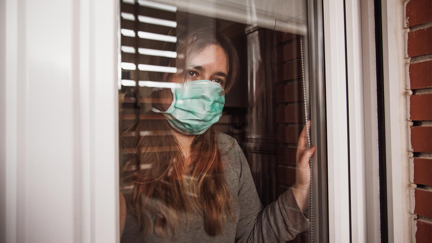 Young woman in quarantine wearing a mask and looking through the window.